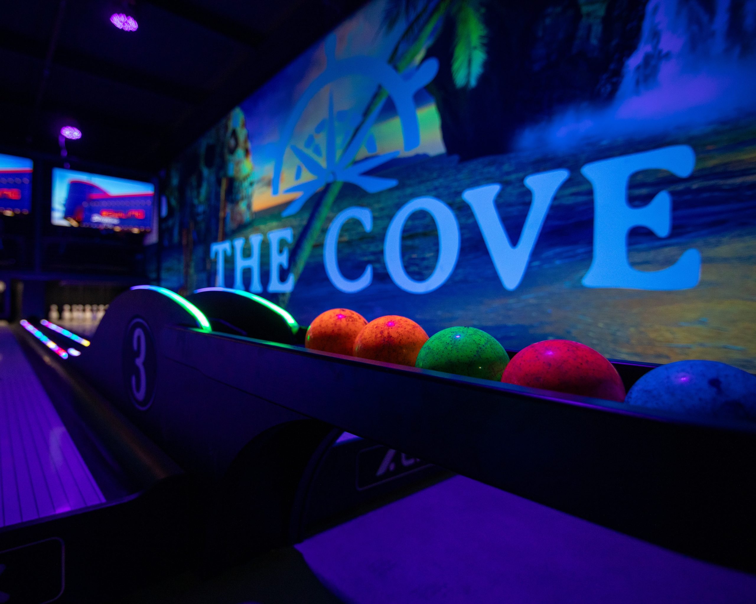 Colorful bowling balls on a return rack under blue and purple neon lights in a bowling alley.