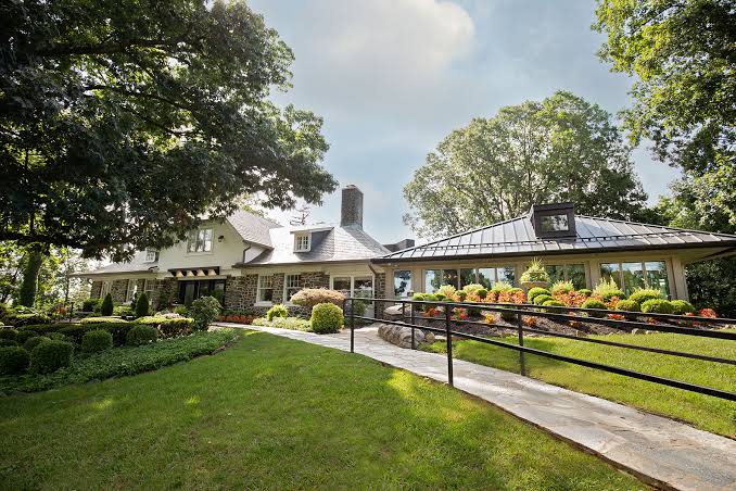 Single-story house with a stone facade, manicured lawn, and shrubs; a curved ramp leads to the entrance.